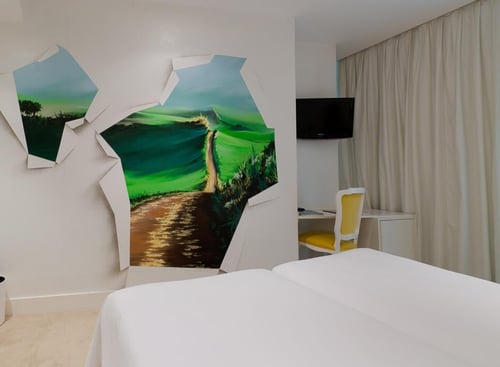 A modern bedroom with a wall featuring a mural of a green landscape and a path, a white bed, a desk with a yellow chair, and