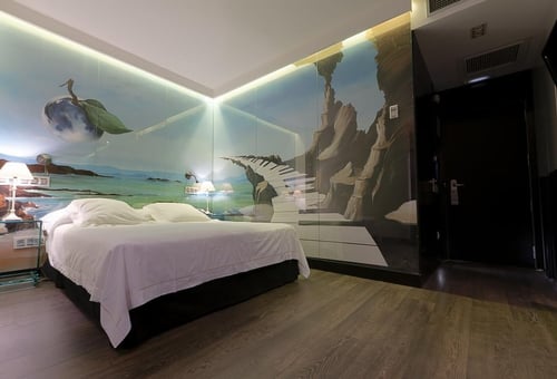 A room with a wall decorated with surrealist murals of landscapes and floating elements, a bed with white sheets, and dark