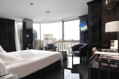 Double room with large panoramic windows, elegant dark-toned furniture, a white bed, and city views. Includes a lounge area