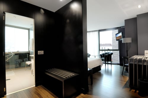 Superior double room with dark walls, a white bed, an armchair next to a panoramic window, and a work area with a desk and a