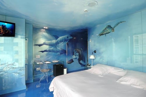 A room with blue walls and ceiling featuring illustrations of dolphins and a sea turtle. It includes a white bed, a desk