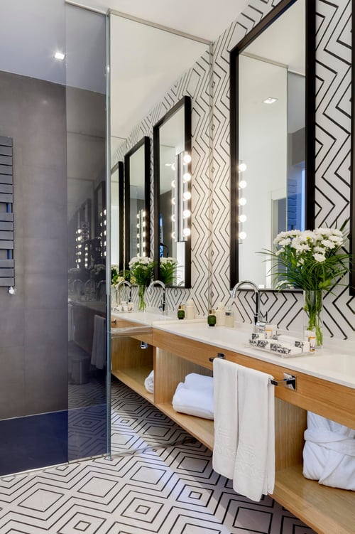 A modern bathroom with black-and-white geometric tiles, a double sink with mirrors, white towels, and warm lighting accents.