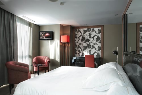 A modern bedroom with a white bed, two red armchairs, a wall-mounted television, and wallpaper featuring leaves in shades of