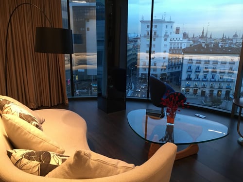 A suite living room with a beige sofa and a glass coffee table, next to large windows offering views of historic buildings