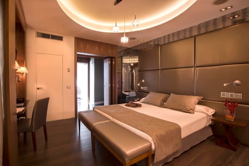 A modern room with a large bed, a padded headboard in shades of gray, warm lighting, and wooden accents. It includes a table