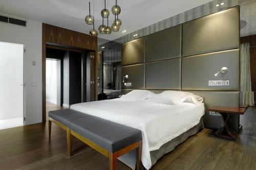 A modern bedroom with a white bed, a gray upholstered headboard, wooden floors, and gold pendant lights. It includes mirrors