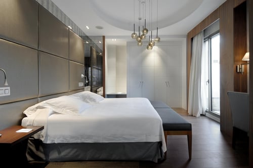A modern room with a large bed, gray padded walls, pendant lights, and dark wood floors. It includes a seating area with a
