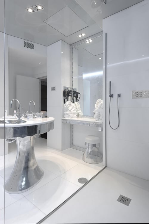 A modern bathroom with a minimalist design in shades of white, featuring a metal sink, mirrors, an open shower, and white