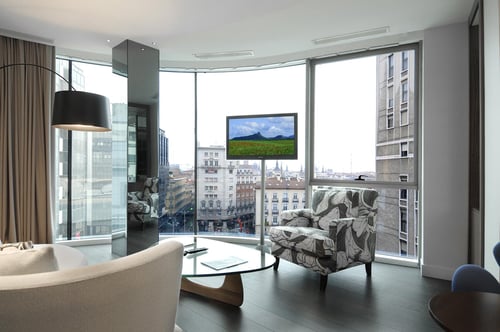 A modern living room with large windows offering city views, light-colored sofas, a floral-patterned armchair, and a
