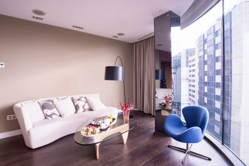 A modern living room with a white sofa, a glass coffee table, a blue chair, and large windows overlooking buildings.