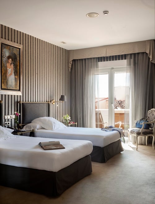 A room with two single beds, gray-and-white vertical stripes on the walls, sheer curtains, a classic painting, and elegant