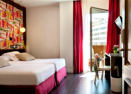 A room with two single beds, light-colored walls, and colorful decor above the headboard. It includes a work area with a