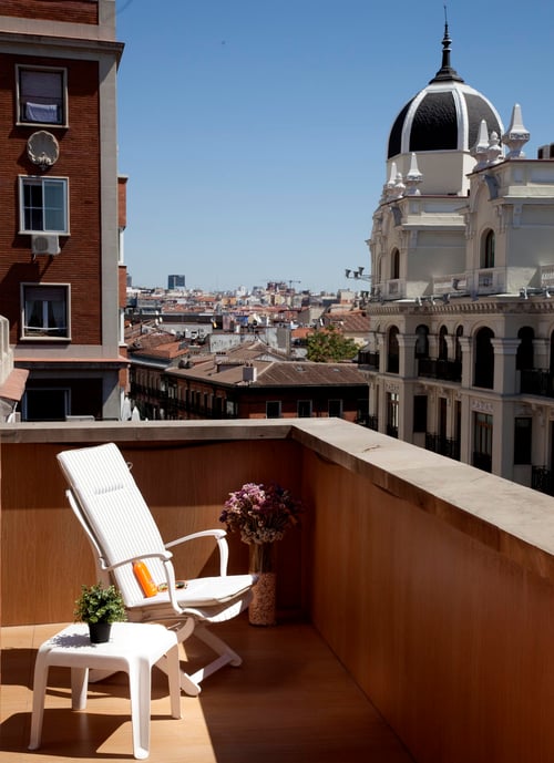 A terrace with a white chair, a side table, and flowerpots, set against historic buildings and a prominent dome on the city