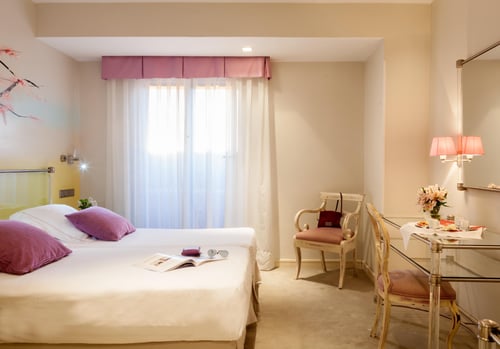 An elegant room with a white bed and purple cushions, a breakfast table, a classic-style chair, and a window with sheer