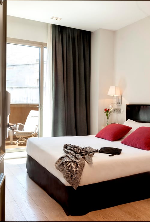 A modern bedroom with a white bed and red cushions, gray curtains, and a balcony with outdoor furniture. Light-colored wood