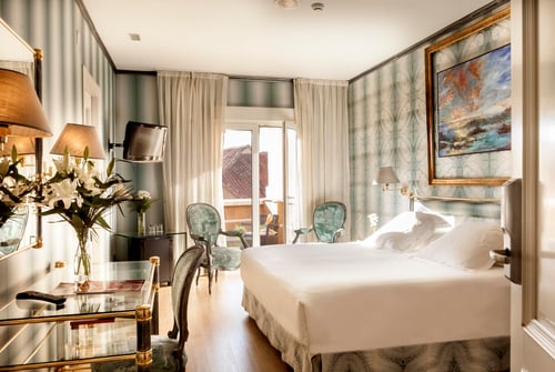 An elegant room with a white bed, light green wallpaper, two designer chairs, tables with lamps, and an abstract painting.