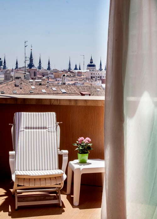 A balcony with a white recliner with gray stripes, a side table with a pot of pink flowers, and a view of an urban skyline