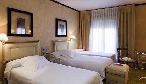 A hotel room with two single beds, beige walls, light-colored curtains, and wall sconces. It includes a painting and a chair