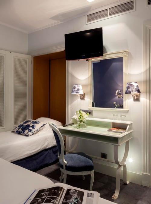 Room with a single bed, a desk with lamps and a mirror, a wall-mounted TV, and a window with curtains. Classic decor in