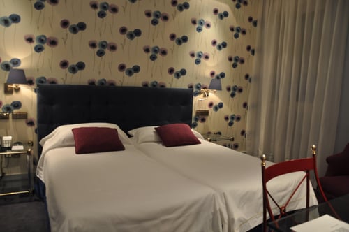 A bedroom with two single beds, a dark headboard and red pillows, a wall with blue and purple floral wallpaper, and wall