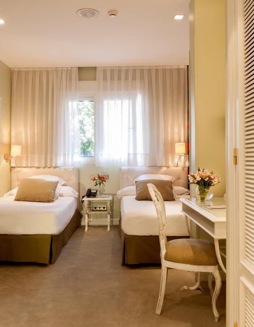 Double room with two single beds, decorated in neutral tones, with white curtains and classic furniture. Includes a table