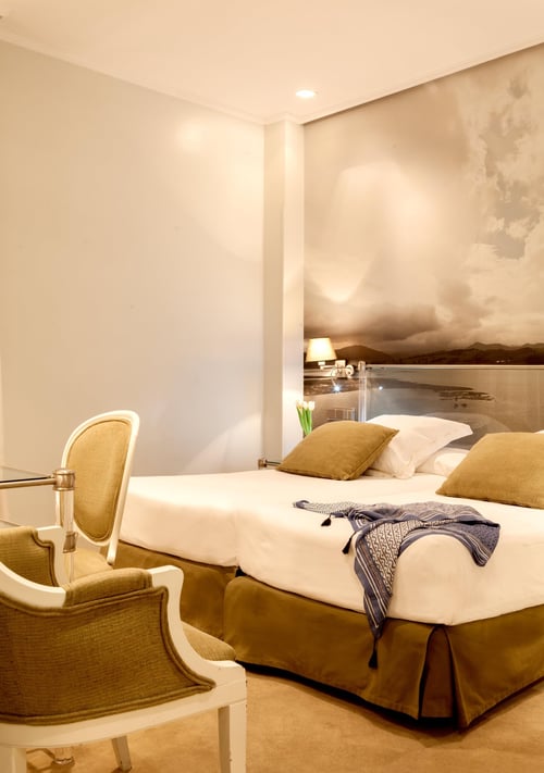 A bedroom with a double bed, beige pillows, and a blue blanket draped over a white comforter. On the wall is a painting of a