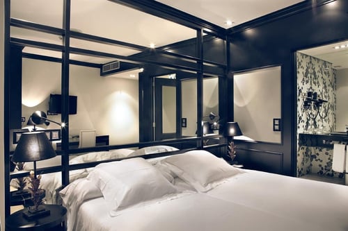 A modern bedroom with a white bed, dark walls, and mirrors that make the space feel larger. It includes table lamps and