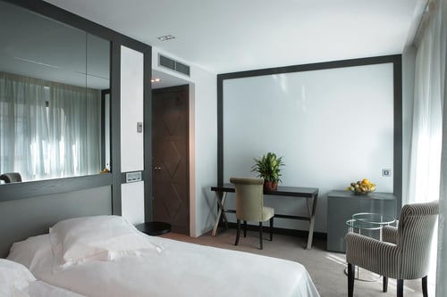 A modern bedroom with a white bed, a desk with a plant and a fruit bowl, light-colored walls, and dark gray accents. It