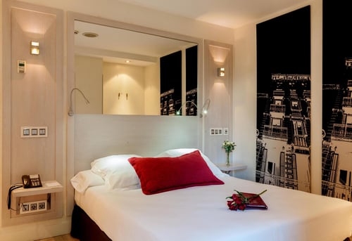 A modern bedroom with a white bed, a red cushion, and a wall featuring a black-and-white urban illustration. It includes