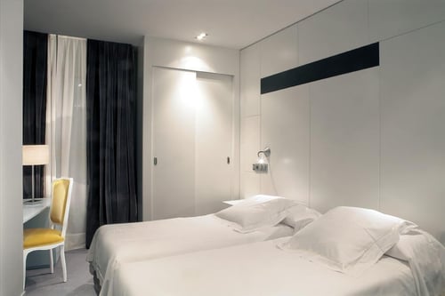 A minimalist bedroom with two single beds, white walls, and a built-in closet. It features a yellow chair, a table lamp, and