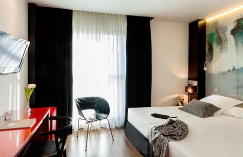 A modern bedroom with a white bed, a black designer chair, a red table, and a TV. Dark curtains and natural light stream in
