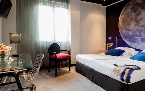 A modern bedroom with a double bed, a glass table, and a designer chair. Highlights include a wall featuring a mural of the