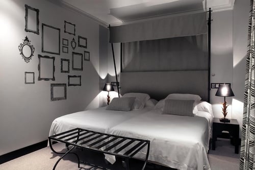 An elegant room with two single beds, gray walls, empty picture frames, table lamps, and a dark mosquito net frame above the