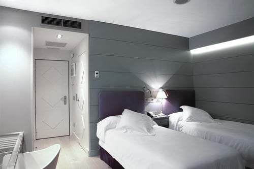 A modern room with two single beds, gray walls, a white door, and a table lamp. It includes air conditioning and ambient