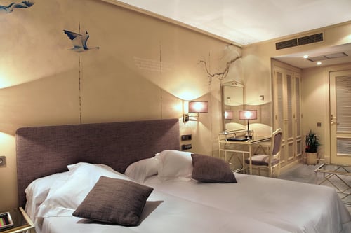 A bedroom with a double bed, beige walls decorated with birds and branches, wall sconces, and a desk area with a mirror and