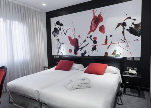 A modern bedroom with a double bed, red cushions, and a wall decorated with abstract art in shades of red, black, and white.
