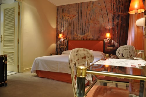 A room with a double bed, a red headboard, and walls decorated with a mural of trees. It includes a glass table with chairs