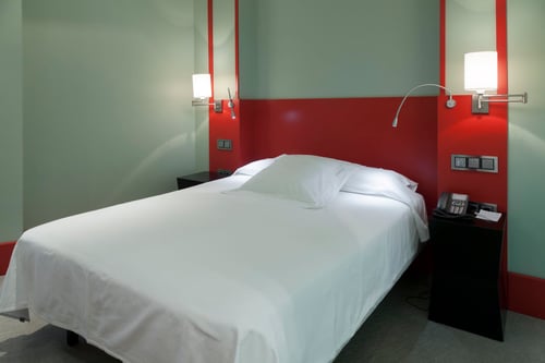 Room with a single bed, walls in shades of light green and red, and adjustable wall lamps. Includes a small table with a
