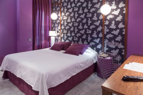 A room with purple walls, wallpaper featuring white hands, and bedding in shades of lilac. Includes a double bed, hanging