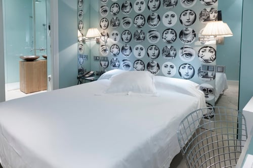 A room with a white bed, a wall decorated with black-and-white images, wall sconces, and a mirror. It includes a metal chair