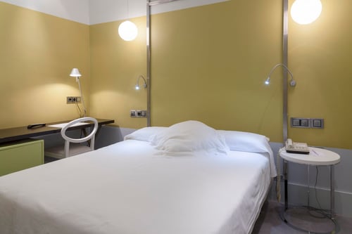 A simple room with light green walls, a single bed with linens, a desk with a chair and a lamp, and a side table with a