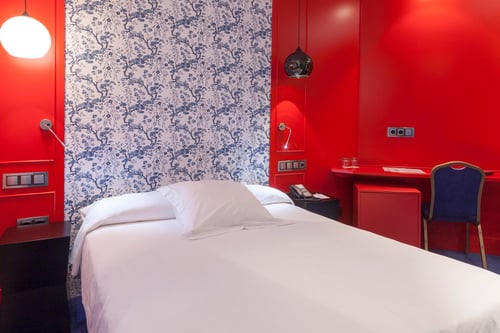 A room with a white bed, red walls, and blue floral wallpaper. It includes a desk, a blue chair, and modern lamps.