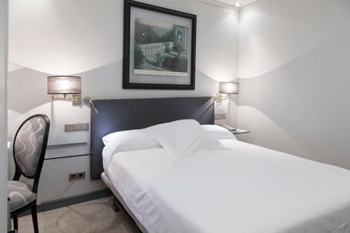 A room with a double bed, light-colored walls, and sconces. It includes a landscape painting, an upholstered chair, and