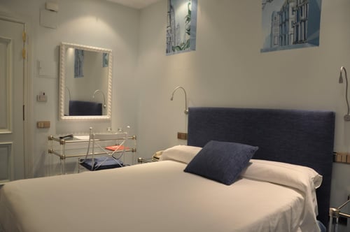 A bedroom with a double bed, a blue headboard, and a matching pillow; light-colored walls with paintings of buildings; and a