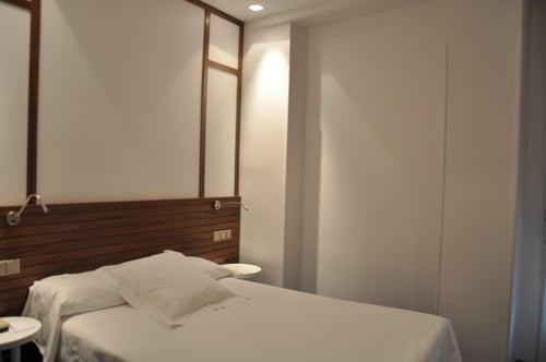 A simple room with a single bed, a dark wood headboard, and white walls. It features recessed lighting and a sliding door.