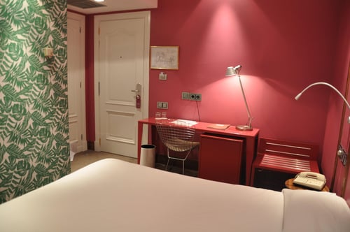 A room with deep red walls and functional furniture, including a red desk, a metal chair, and a table lamp. Details include