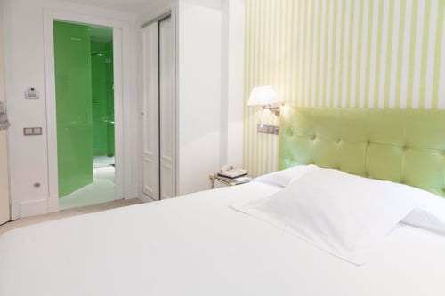 A modern bedroom with a white bed, a light green padded headboard, and a wall with yellow and white vertical stripes. A