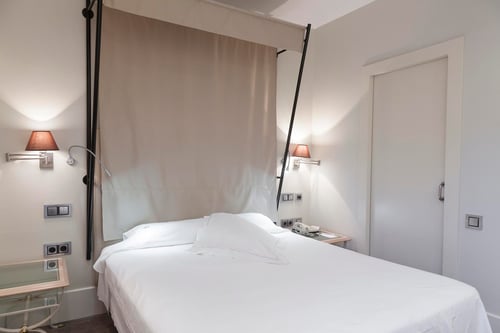 A minimalist bedroom with a white bed, light-colored walls, and wall sconces. It includes a white door, a beige curtain, and