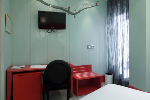 A room with light-colored walls and a tree-and-birds motif, red furniture, a desk, a black chair, a television, and a dark